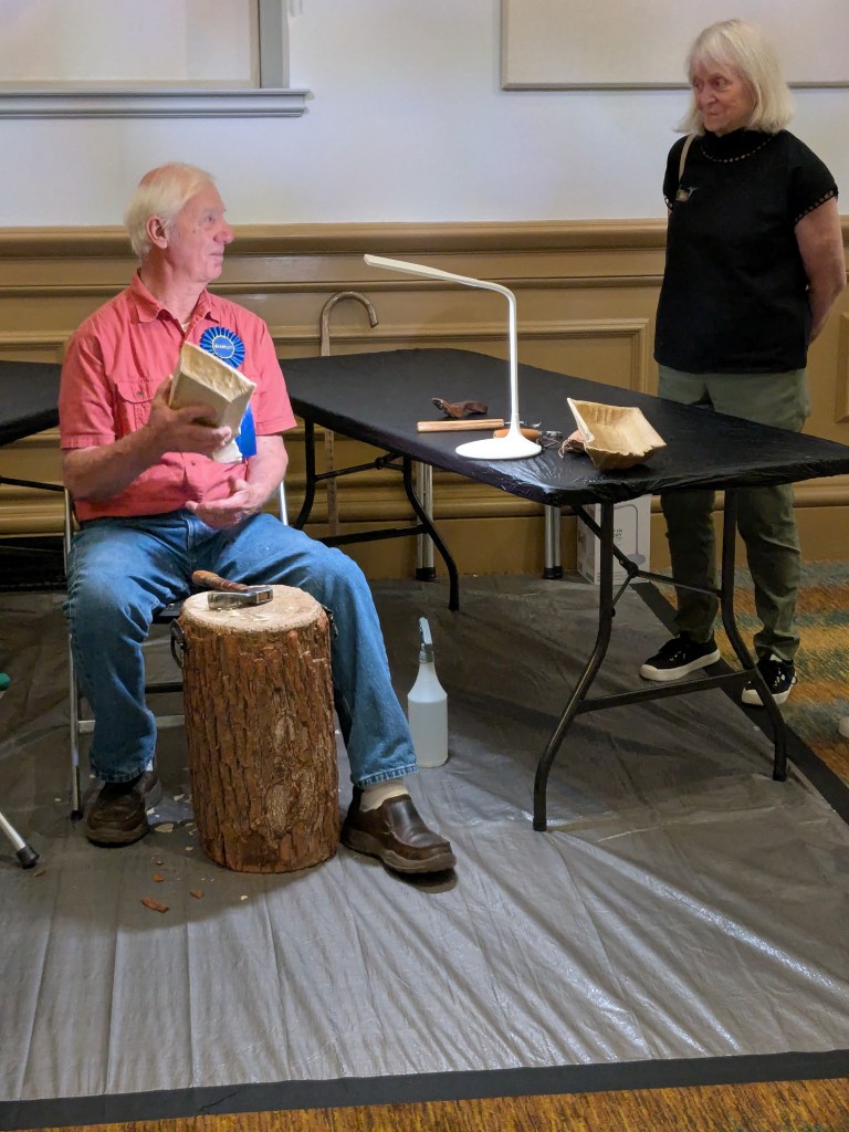 A man uses tools for carving on a wooden stump, used as a bench. Indoors, inside a museum.