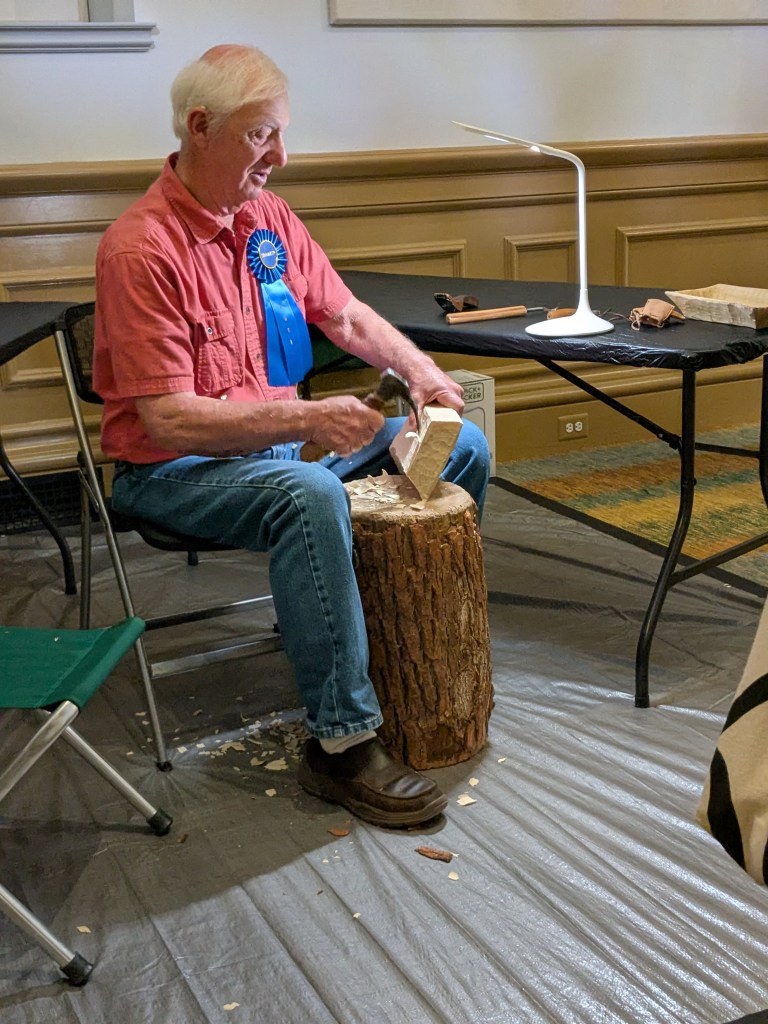 A man uses tools for carving on a wooden stump, used as a bench. Indoors, inside a museum.