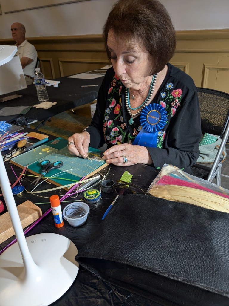 A woman seated is doing delicate craftwork on a table with craft materials nearby.