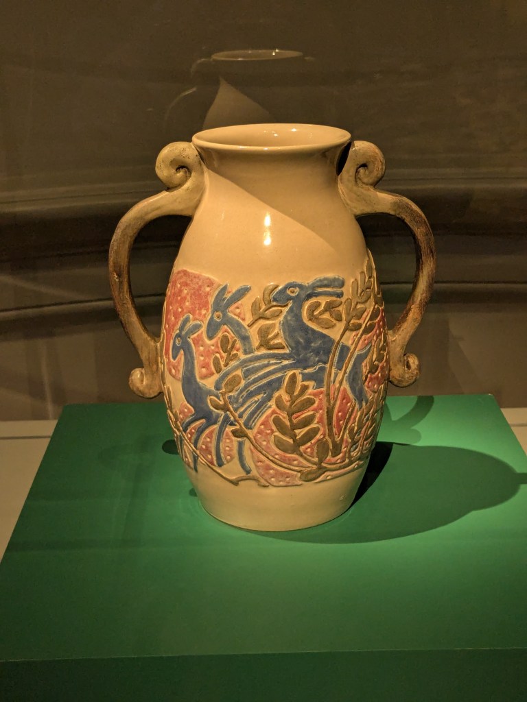A ceramic vessel of tan pottery with colored designs and two handles on opposite sides.