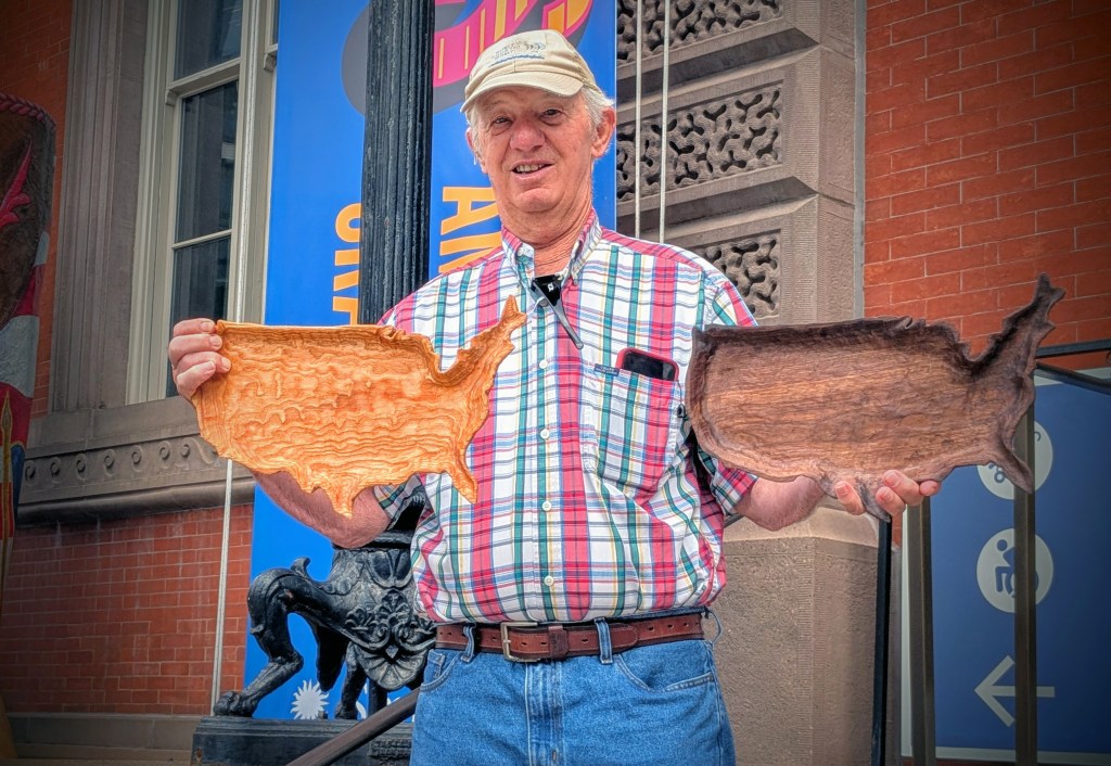 A man holding two carved wooden trays shaped like a map of the continental USA outside a museum building.